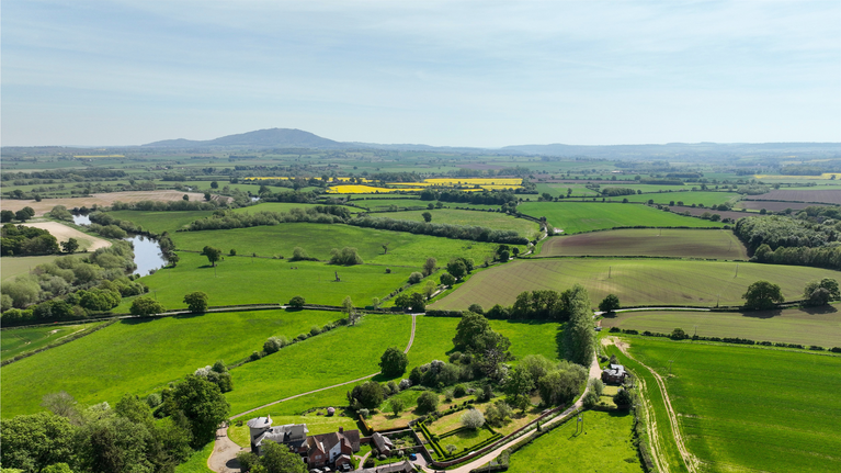 Drone image showcasing the agricultural landscape at Attingham Park, Shropshire, with green fields, winding hedgerows, a river meandering through the scene, and distant hills under a clear blue sky.